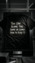 Black t-shirt with bold white motivational typography reading 'You Can Blame the Game or Learn How to Play It'